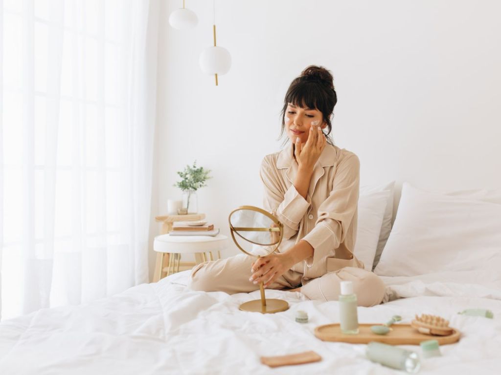 woman sitting on bed with natural skin care products enjoying some self-care. 